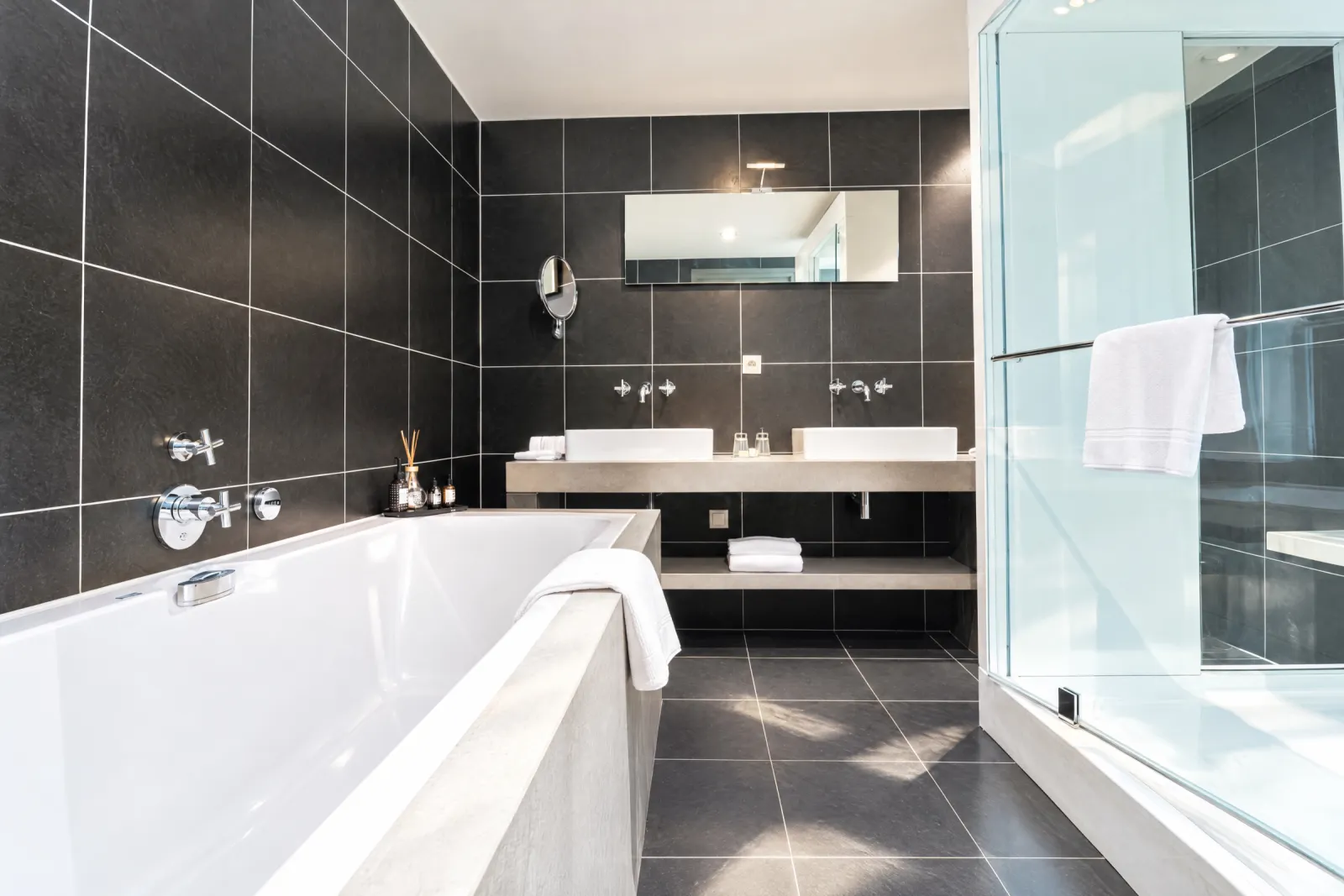 Badkamer van de Superior Suite met zwarte tegels, ligbad, dubbele wastafel en douche